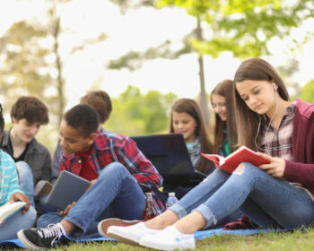 Pre-teenage and teenage group of boys and girls studying, hanging out together in local park or school campus with friends.  Teen girl reading in foreground.